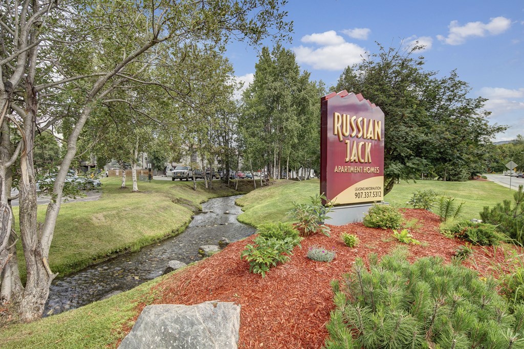 Sign for Russian Jack Apartment Homes by a small creek with red mulch landscaping, surrounded by lush trees and a clear blue sky, creating a peaceful setting.