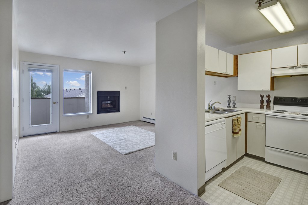 Cozy apartment interior here at Silver Ridge Apartments with carpeted living room, fireplace, and bright window. Adjacent kitchen has white appliances and cabinets, and a warm tone.