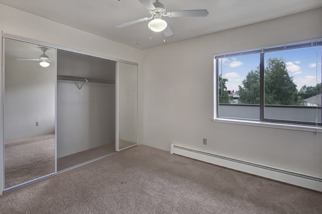 Empty bedroom here at Silver Ridge Apartments with beige carpet and white walls. Large mirrored closet doors reflect a ceiling fan. Bright window view reveals green trees and blue sky.
