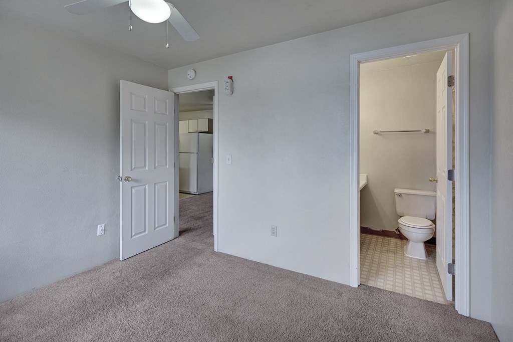 Empty bedroom here at Silver Ridge Apartments with gray walls and carpet, open door to a bathroom with a toilet and towel rack. Ceiling fan overhead. Doorway reveals a kitchen area with a fridge.