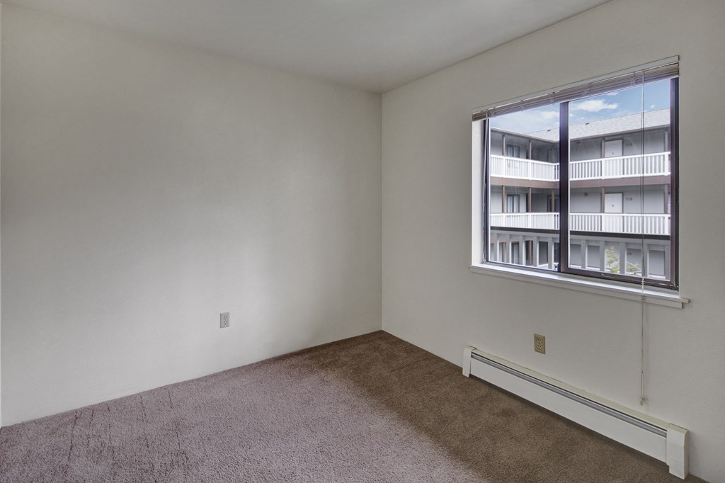 Empty bedroom here at Silver Ridge Apartments with beige carpet, white walls, and a window showing an apartment building courtyard. The light creates a calm, neutral tone.