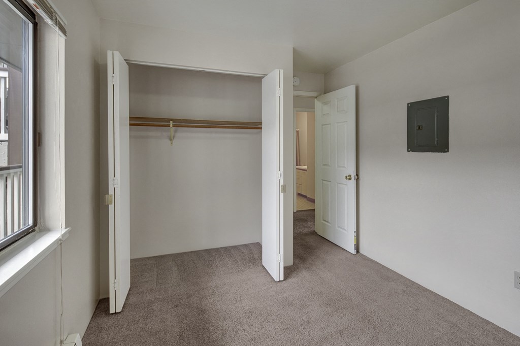 Empty bedroom here at Silver Ridge Apartments with beige carpet, open closet with wooden rod, white walls, and doors leading to another area. A window on the left brings in light.