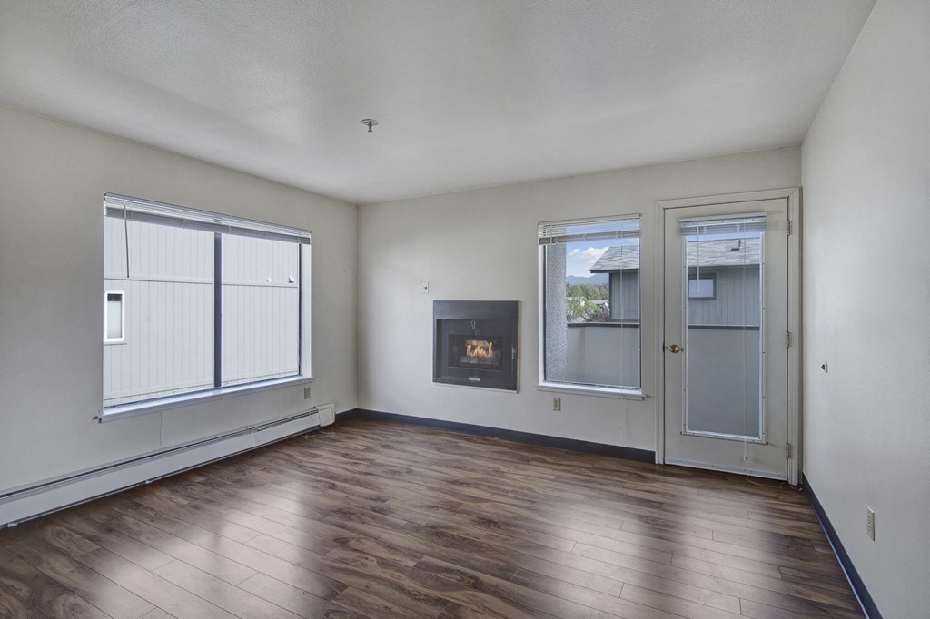 Empty living room here at Silver Ridge Apartments with light wood flooring, white walls, and a modern fireplace. Large windows and a glass door provide natural light. Cozy and minimalistic.