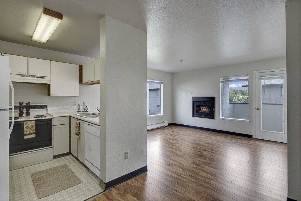 A small kitchen here at Silver Ridge Apartments with light cabinets, white appliances, and a cozy rug opens into a bright living room with hardwood floors and a corner fireplace.