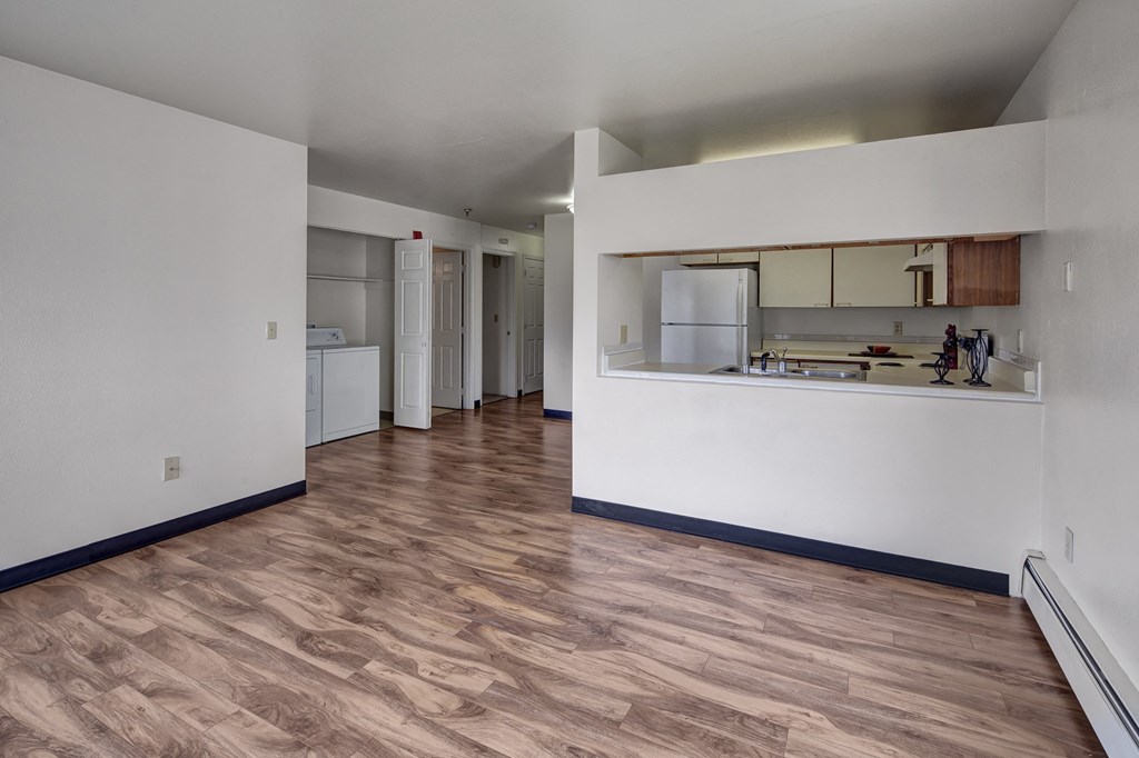 Spacious, empty living room here at SIlver Ridge Apartments with light wood flooring and white walls. Open kitchen with a visible fridge and countertop, providing a clean, minimalist feel.