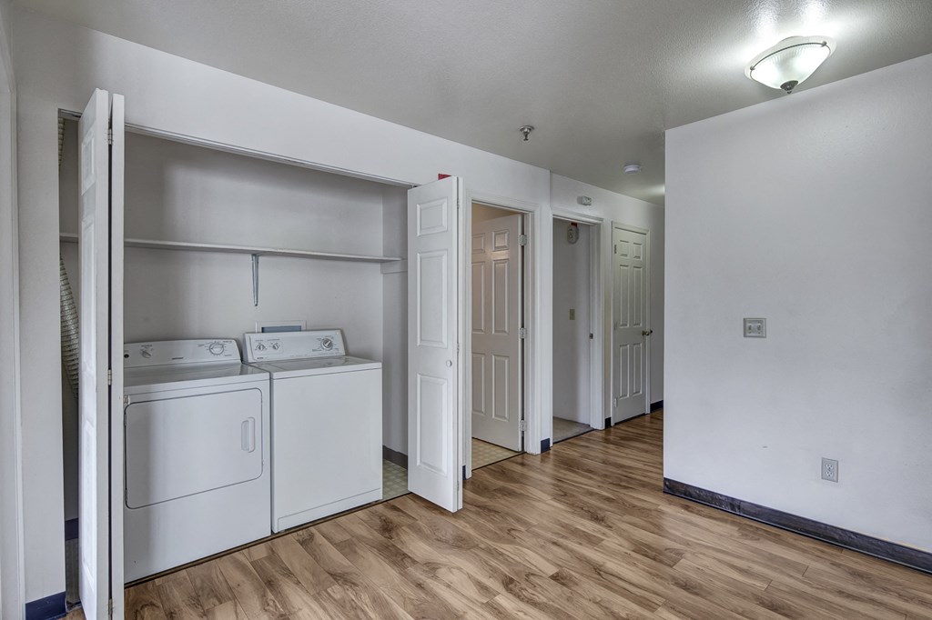 Bright laundry area here at Silver Ridge Apartments with white washer and dryer set against a light wall. Wood flooring leads to open doors revealing more rooms, under soft overhead lighting.