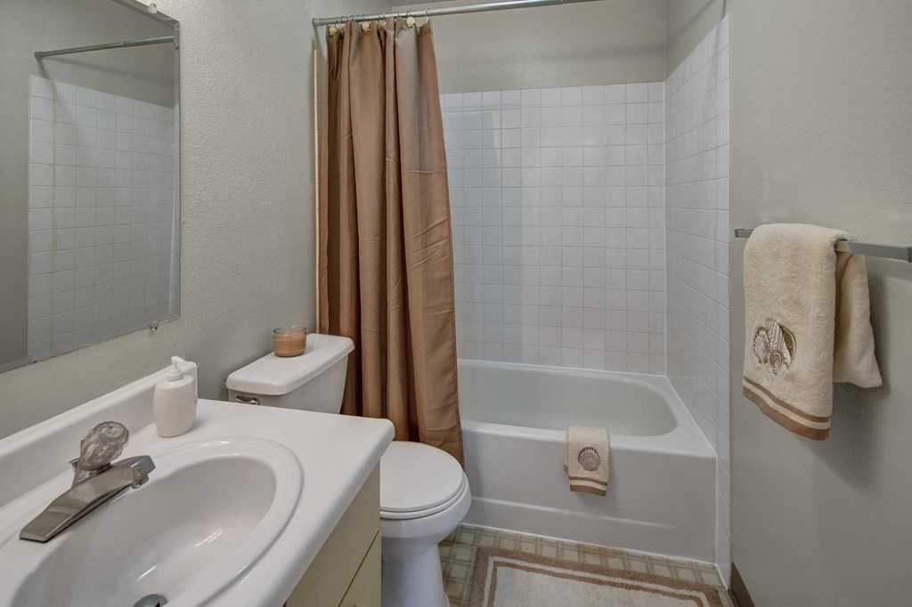A small bathroom here at Silver Ridge Apartments with white sink and toilet, beige shower curtain, and matching towels with shell motifs. A candle adds a warm, inviting touch.