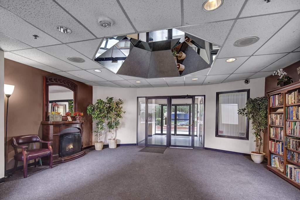 Lobby here at Silver Ridge Apartments with a cozy and inviting feel, featuring a dark wooden fireplace with a mirror above it, two potted plants, a bookshelf full of books, and seating.