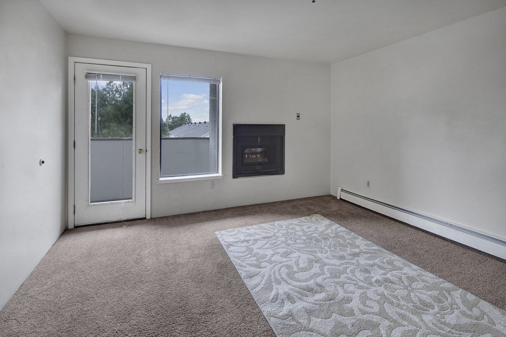 Empty living room here at Silver Ridge Apartments with white walls, a glass door, and a window showing trees outside. A fireplace is set into the wall, and a carpet with floral patterns lies on the floor.