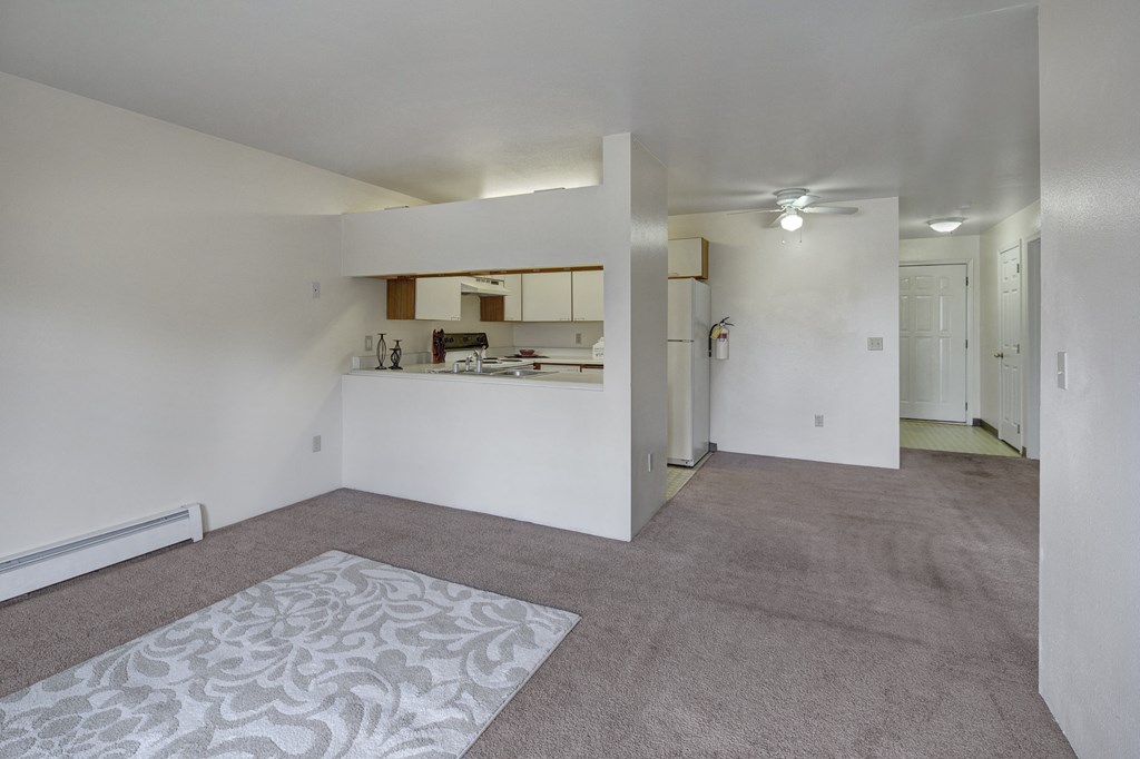 Spacious living room here at Silver Ridge Apartments with beige carpet and patterned rug. Open view of a white kitchen with appliances. Ceiling fan and soft lighting create a cozy atmosphere.