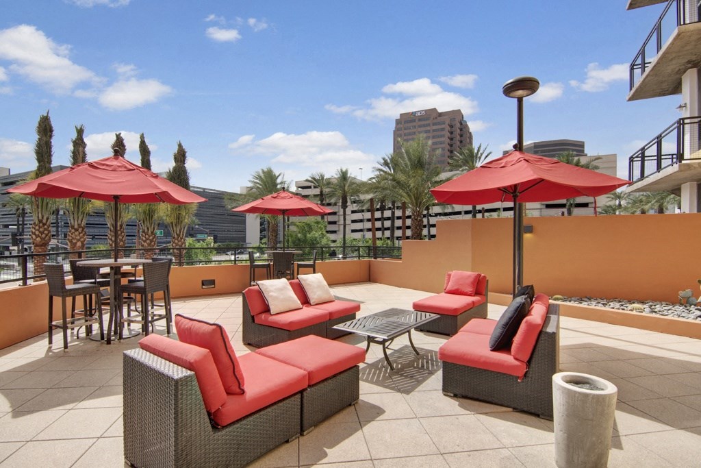 Outdoor patio here at Skyline Lofts Apartments with red cushioned wicker furniture and red umbrellas under a clear blue sky. Surrounded by palm trees and city buildings, conveying a relaxed urban vibe.