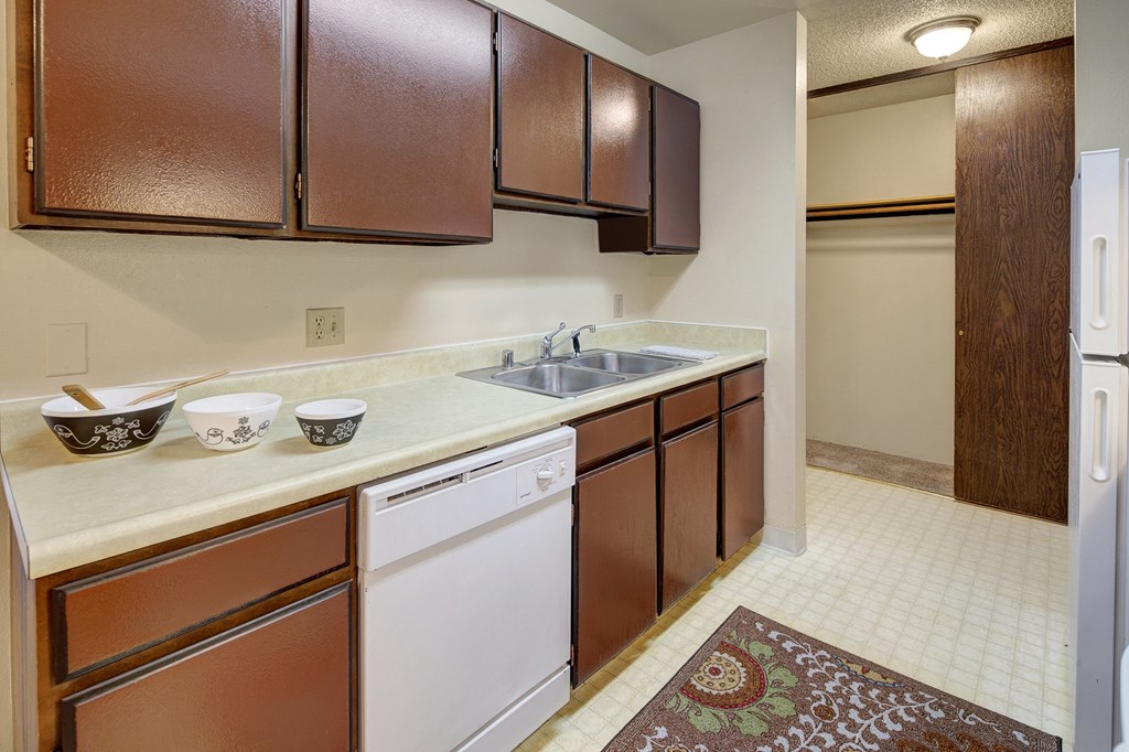 A kitchen here at Taiga Apartments with dark brown cabinets and a light countertop. Three decorative bowls and a wooden spoon sit by the sink. There's a patterned rug on the beige floor.