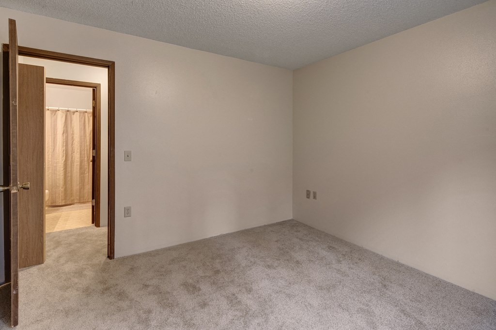 Empty beige carpeted bedroom here at Taiga Apartments with a doorway to a bathroom, partially visible beige shower curtain. Neutral tones convey a calm, minimalist atmosphere.