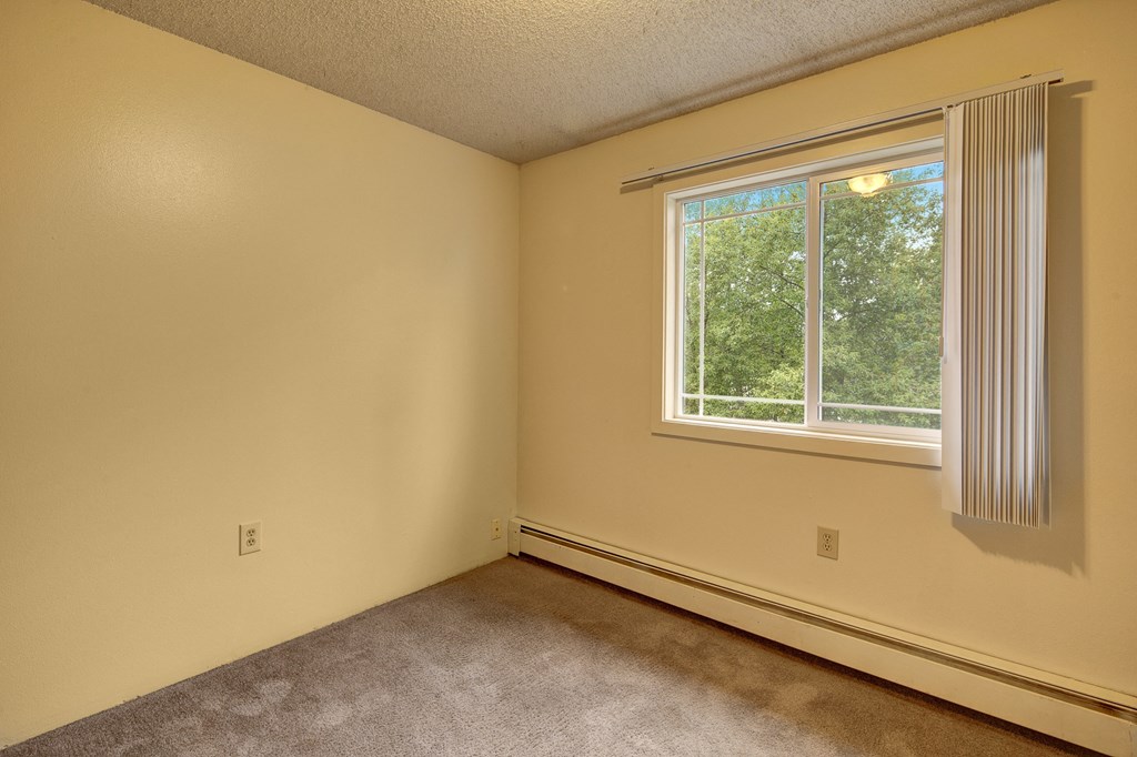 Empty bedroom here at Taiga Apartments with beige walls, carpeted floor, and a large window with vertical blinds. The window reveals lush green trees outside, conveying tranquility.
