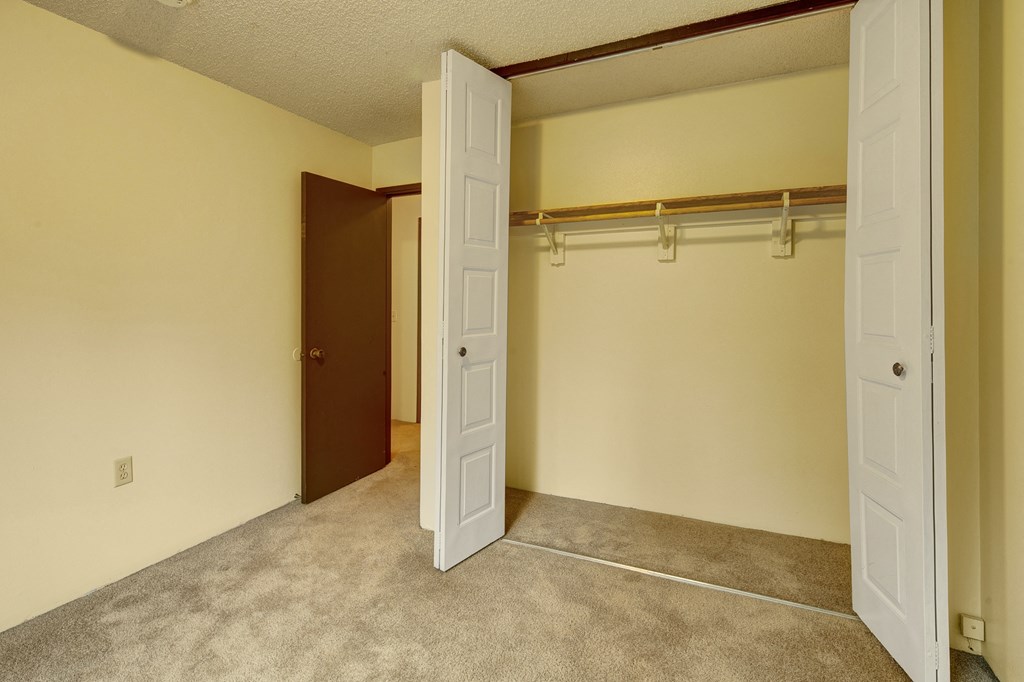 Empty beige room here at Taiga Apartments with open closet doors, revealing a wooden rod and shelf inside. Carpeted floor and an open brown door leading to another room. Quiet and clean atmosphere.