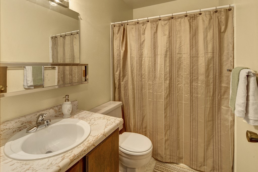 A small bathroom here at Taiga Apartments with a marble-patterned sink, soap dispenser, and toothbrush holder. Beige shower curtain, wooden door with towels, bright lighting.
