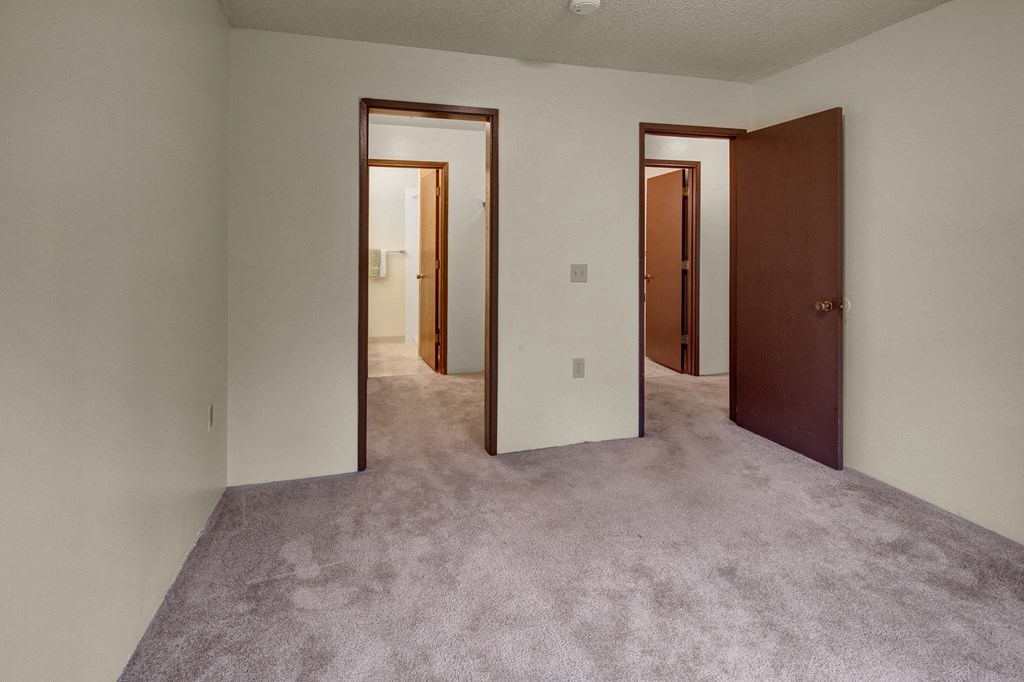 Empty bedroom here at Taiga Apartments with beige carpet, white walls, and open brown doors leading to two additional rooms. The atmosphere is neutral and minimalistic.