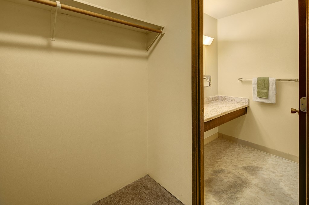 Minimalist interior here at Taiga Apartments showing an empty closet with a wooden rod and shelf. Adjacent bathroom features a marble countertop and towel rack with towels.