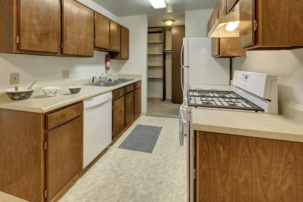 Narrow kitchen here at Taiga Apartments with wooden cabinets, beige countertops, a gas stove, white appliances, and a sink. A pantry, grey rug, and scattered bowls create a cozy feel.