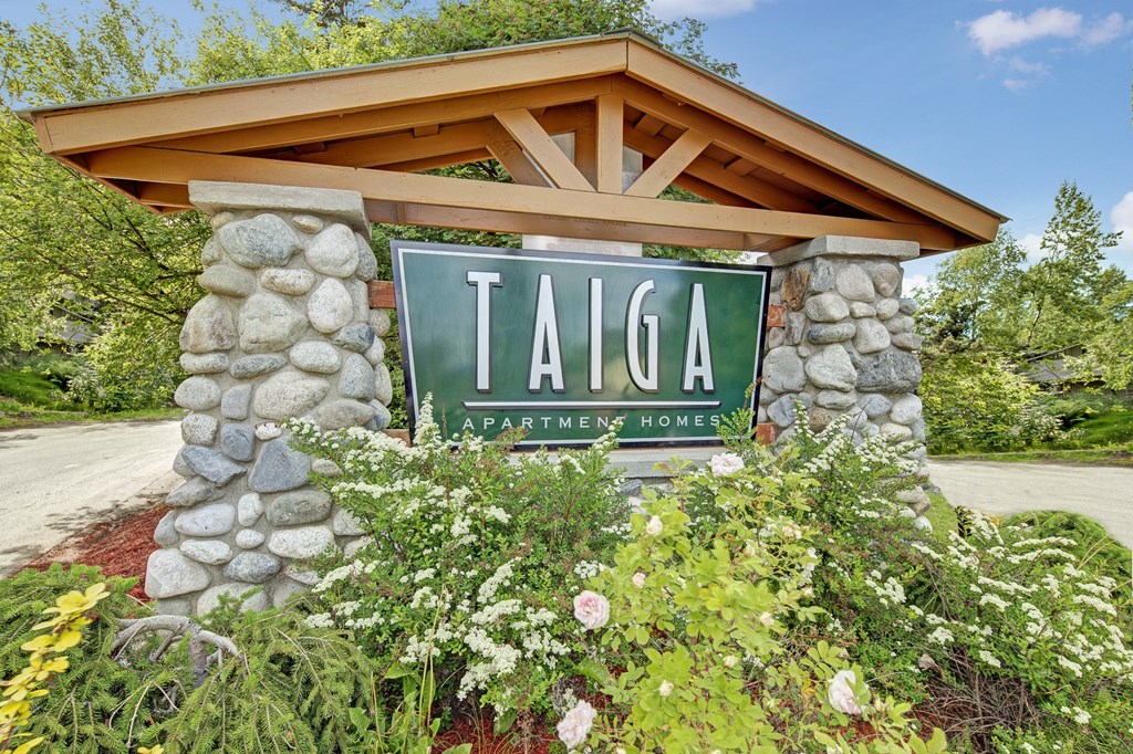 Stone sign for Taiga Apartment Homes amid lush green foliage and blooming flowers, set against a bright blue sky, conveying a welcoming and serene atmosphere.