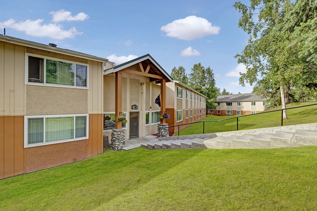 Two-story Taiga Apartment building with beige and orange siding, large windows, and a path leading up to the entrance. Lush green lawn and trees surround it.