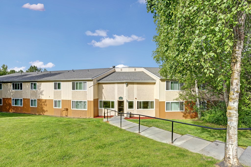 Two-story beige Taiga Apartment building with green-trimmed windows under a clear blue sky. A tree and lush grass flank the entrance, creating a serene atmosphere.