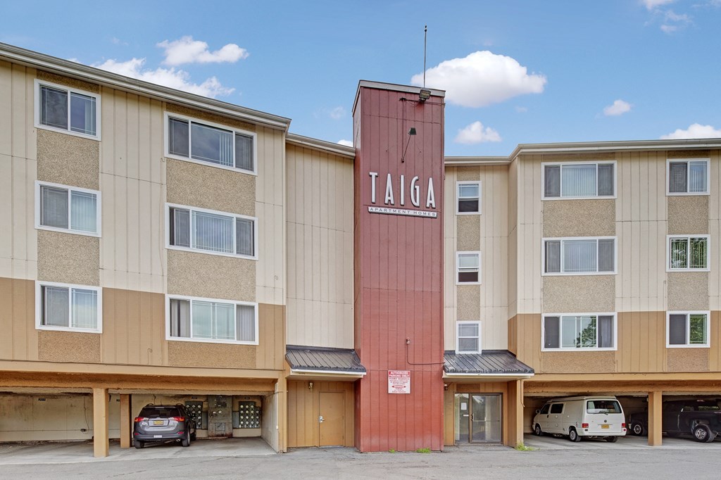 Four-story Taiga Apartment building with a vertical red sign reading "TAIGA." Beige facade with multiple windows and cars parked in ground floor garages.