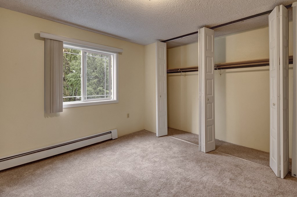 Empty carpeted bedroom here at Taiga Apartments with a large open closet featuring white folding doors. A window with blinds is on the left, revealing greenery outside. Neutral tones.