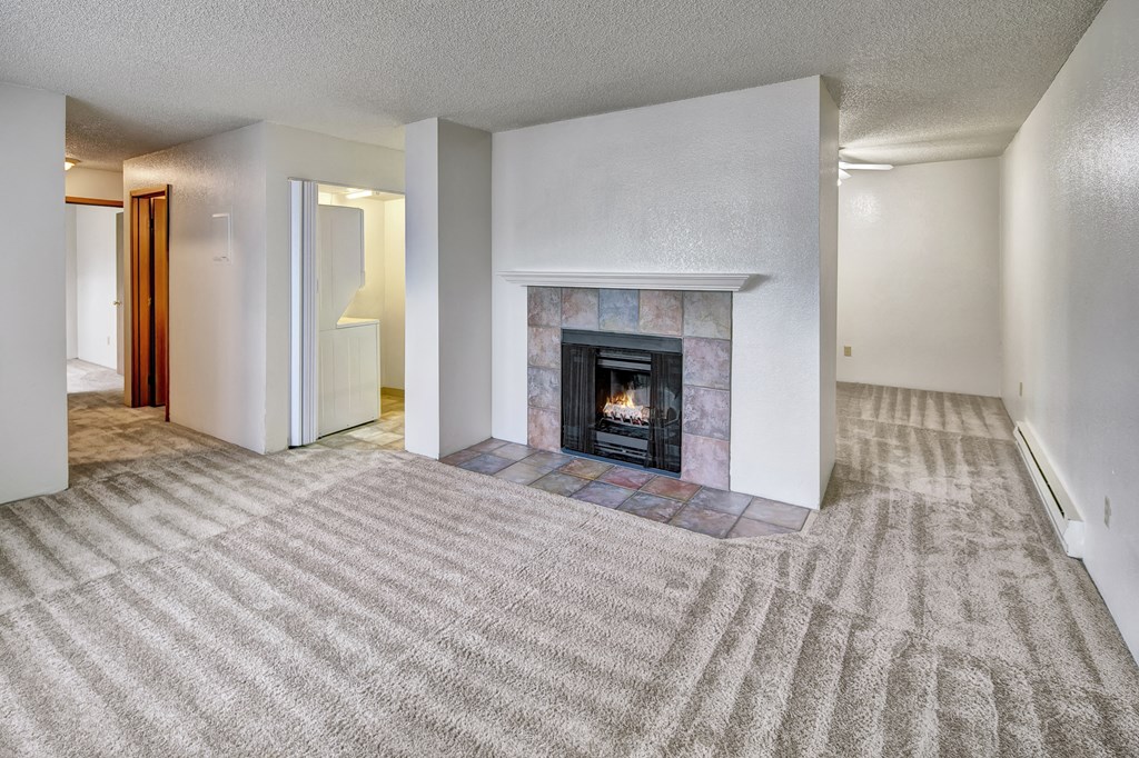 A spacious living room here at Terrace on the Lake Apartments with beige carpet features a lit fireplace with a tiled mantel. The room is bright, with white walls and a cozy feel.