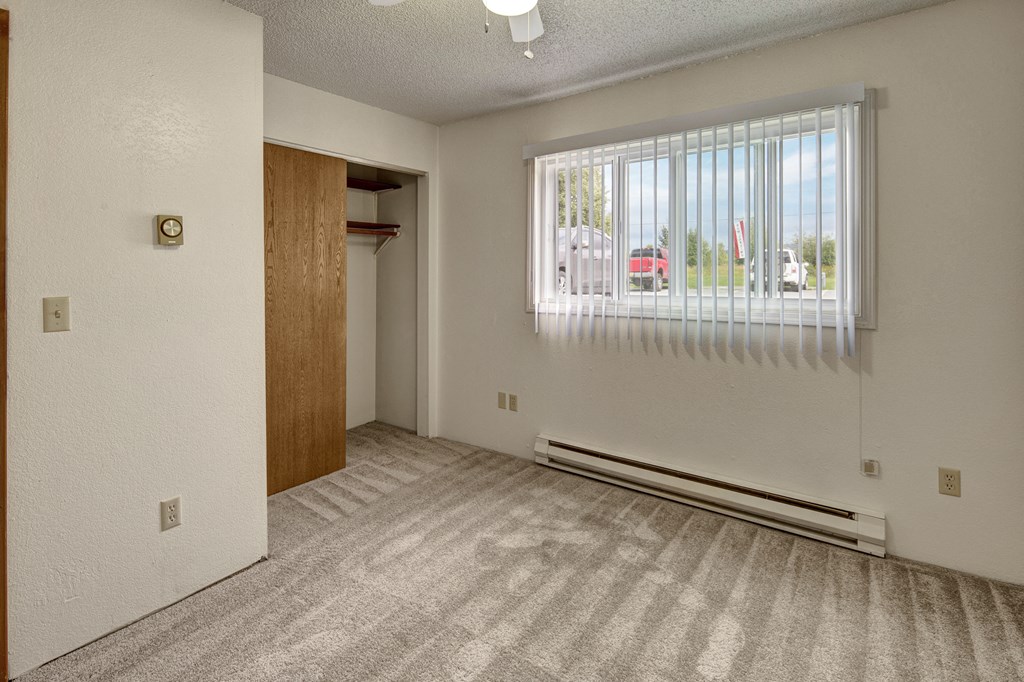 Empty bedroom Terrace on the Lake Apartments with beige carpet, white walls, and vertical blinds on a window. An open closet with a wooden door is on the left, creating a neutral, clean atmosphere.