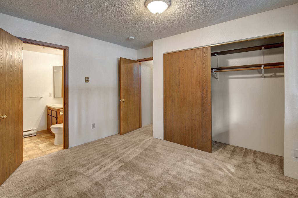 Empty bedroom here at Terrace on the Lake Apartments with beige carpet, a wooden closet with sliding doors, and an open door revealing a bathroom. Soft lighting creates a neutral, calm tone.