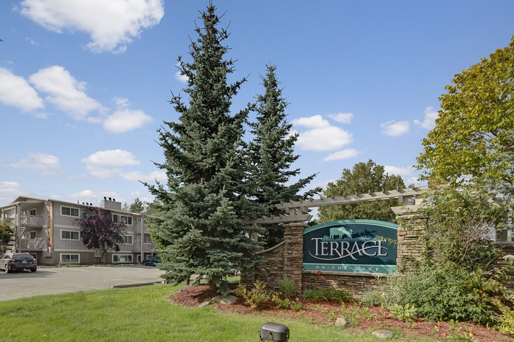 Apartment complex entrance with stone sign reading "Terrace on the Lake," flanked by tall evergreen trees. A building and parked cars are visible under a bright blue sky.