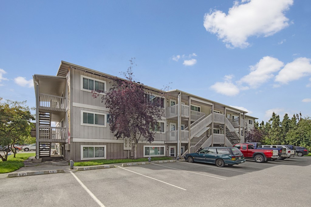 Three-story beige Terrace on the Lake Apartment building with external staircases, parked cars in front, and a cloudy blue sky. Trees frame the scene, offering a serene feel.