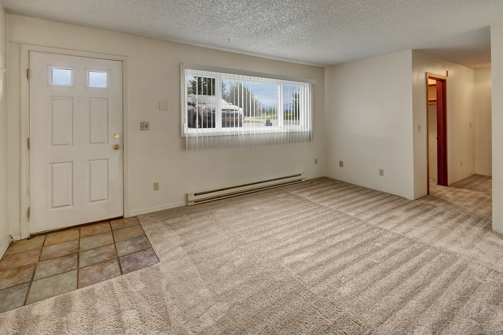 Empty living room here at Terrace on the Lake Apartments with beige carpet, white walls, and a light blue door with windows at the top. Large window with vertical blinds lets in natural light.