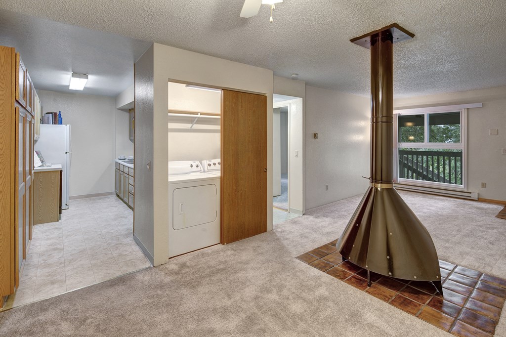Open-plan living space here at Terrace on the Lake Apartments with beige carpet, a central vintage cone fireplace, and a windowed door leading to a balcony. Adjacent is a utility room with a washer and dryer.