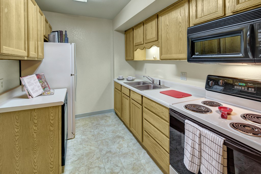 A narrow kitchen here at Terrace on the Lake Apartments with wooden cabinets, a double sink, stove, and microwave. A red cutting board and striped towels add a cozy touch. Bright lighting.