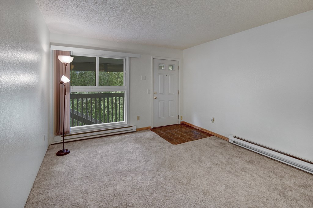 Empty bedroom here at Terrace on the Lake Apartments with beige carpet, white walls, and a ceiling light. A large window with a view of trees is on the left, next to a white door.