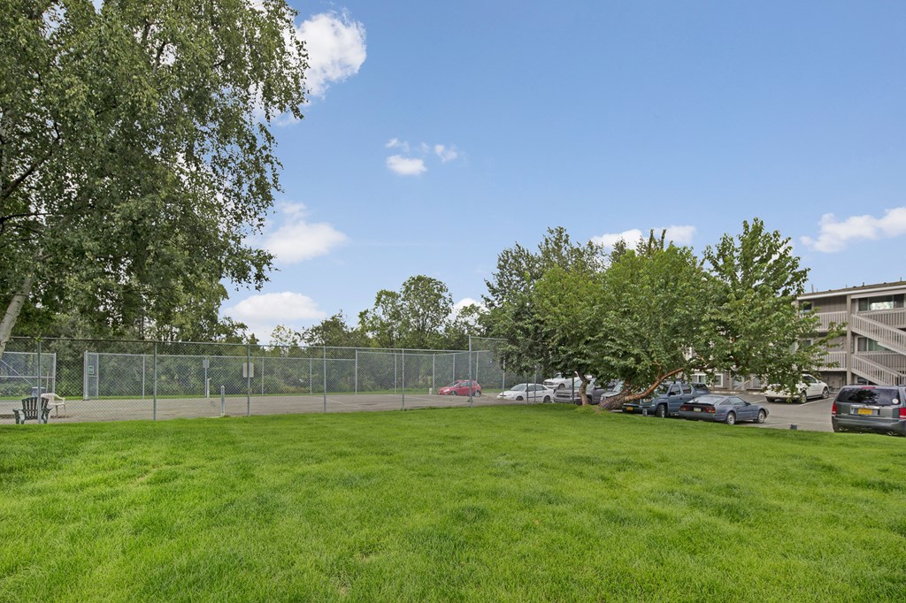 Lush green lawn in the foreground, fenced tennis court to the left, and parked cars to the right. Clear blue sky and leafy trees create a serene atmosphere here at Terrace on the Lake Apartments.