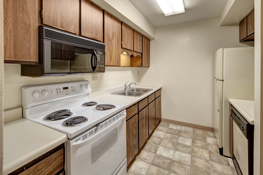 Narrow kitchen here at Terrace on the Lake Apartments with brown wood cabinets, white appliances, and stone-patterned linoleum floor. Features a stove, microwave, sink, and fridge.