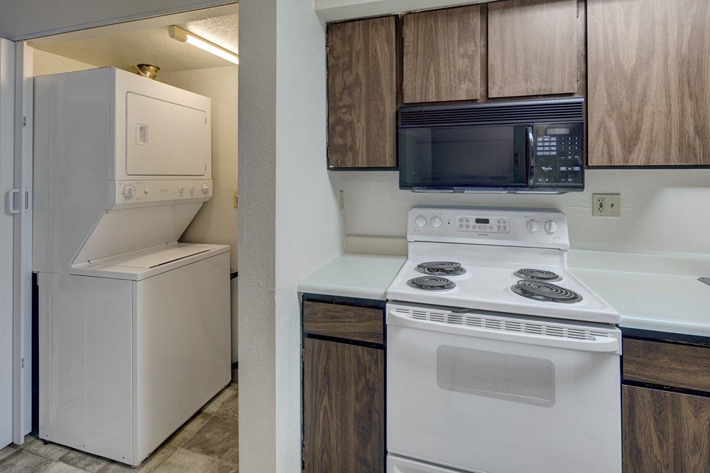 Compact kitchen here at Terrace on the Lake Apartments with white stove, dark wood cabinets, and built-in microwave. Adjacent, a stacked washer and dryer in a small laundry nook.