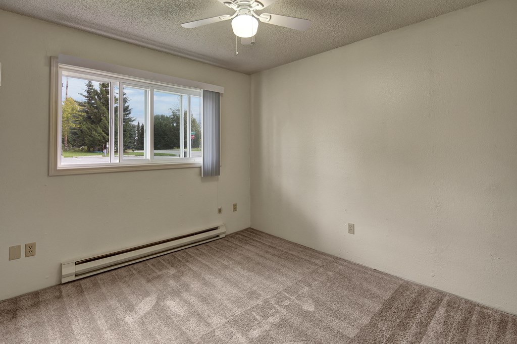Empty bedroom here at Terrace on the Lake Apartments with beige carpet and pale walls, featuring a large window with venetian blinds. A ceiling fan hangs above, and sunlight creates gentle shadows.
