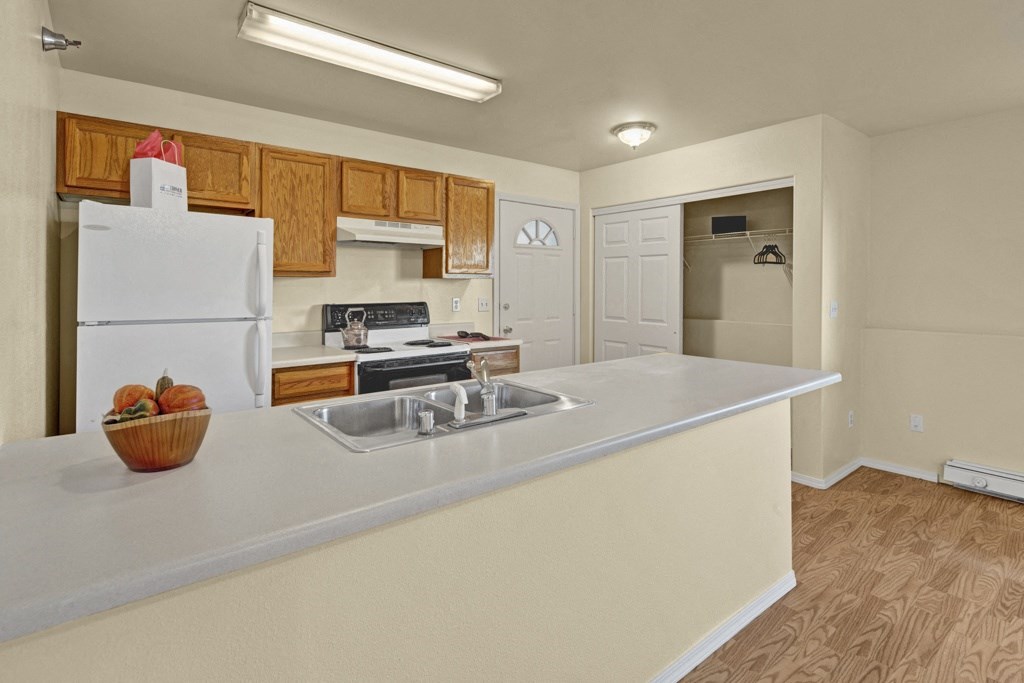 Kitchen here at Timber Ridge Apartments with wooden cabinets, white appliances, and a countertop with a sink. A bowl of squash is on the counter. Closet with hangers is open.