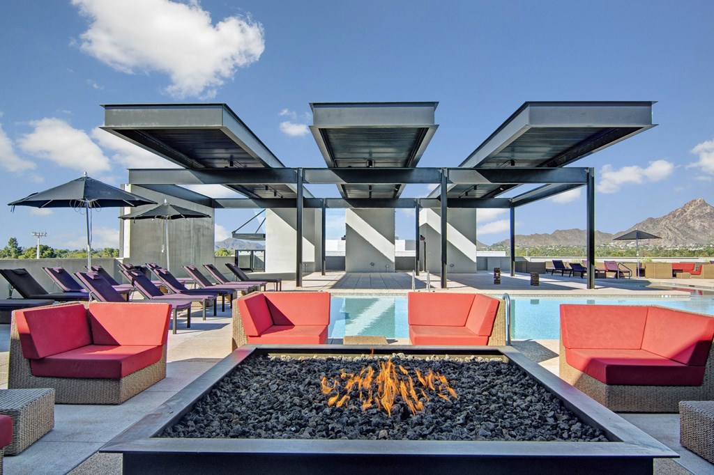 Rooftop pool here at Trend at 51 Apartments with a modern fire pit surrounded by red chairs. Purple lounge chairs and black umbrellas line the deck. Mountains and blue sky in the background.