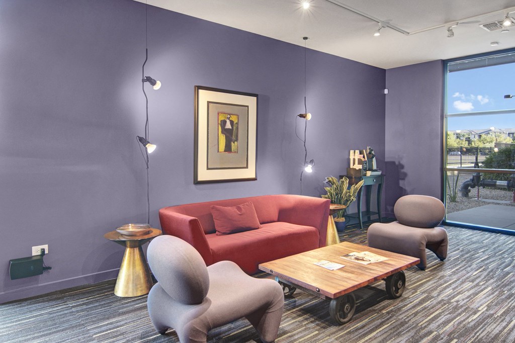 Modern lounge here at Trend at 51 Apartments with a purple accent wall, a red sofa, abstract artwork, quirky gray chairs, and a wooden table. Warm, inviting ambiance.
