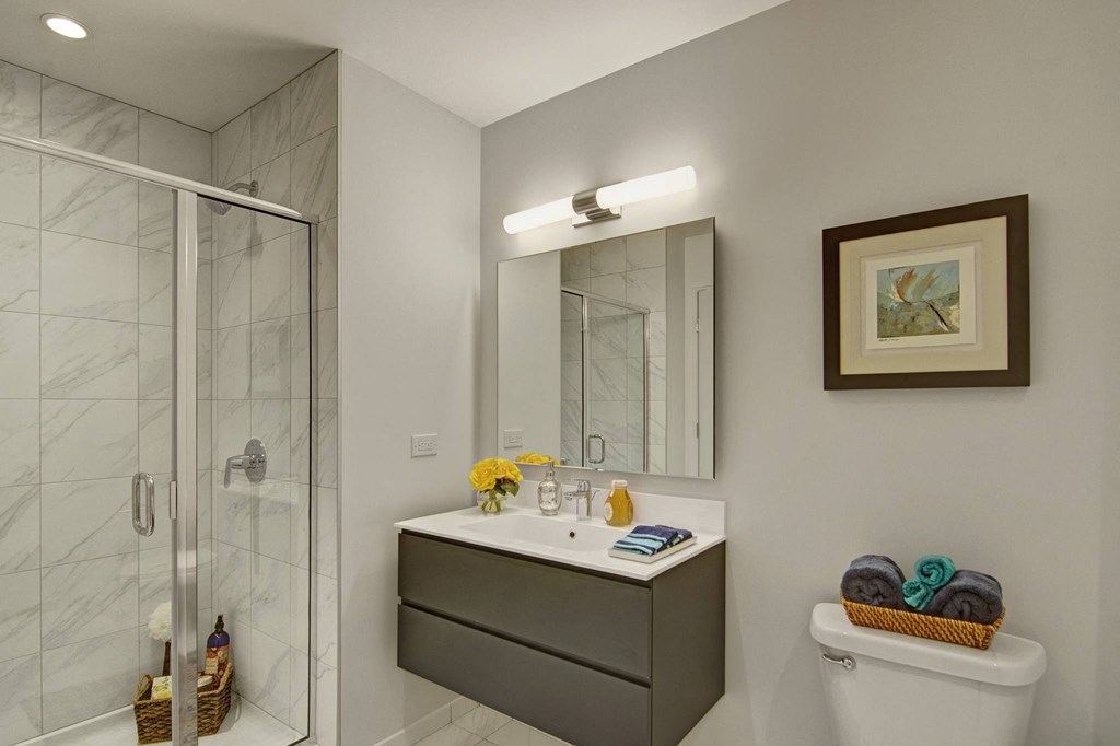 Modern bathroom here at Trend at 51 Apartments with a sleek vanity, large mirror, and bright lighting. A glass shower is on the left. A basket of towels sits atop the toilet, adding a cozy touch.
