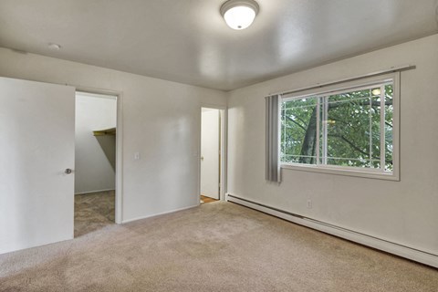 An empty bedroom here at the Village at Calais Apartments with beige carpet, white walls, and a window with vertical blinds showing green foliage outside. Open doors reveal a closet and another room.