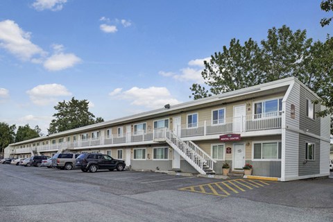 Two-story Village at Calais Apartment building with a staircase in the center, surrounded by parked cars. Clear blue sky, trees in the background; welcoming, calm atmosphere.