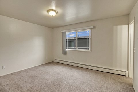 Empty bedroom here at the Village at Calais Apartments with beige carpet and off-white walls, featuring a large window with blinds and a ceiling light. Natural light creates a bright, neutral ambiance.