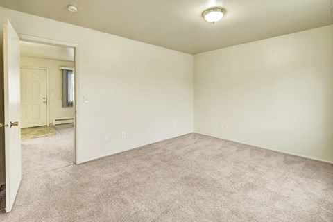 Village at CalaisEmpty beige room here at the Village at Calais Apartments with carpeted floor, plain walls, and an overhead light fixture. An open door reveals an adjacent space with a window and a closed front door. Minimalist and calm ambiance. Apartments - Livingroom