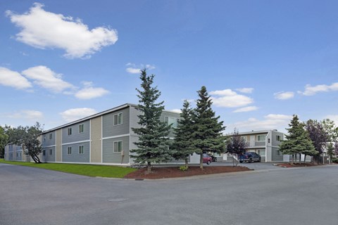 Two-story gray Village at Calais Apartment buildings with white trim, surrounded by lush trees and neatly manicured lawns under a bright blue sky with scattered clouds.